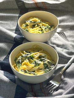 two white bowls filled with pasta and spinach on top of a bed next to a fork