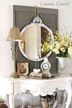 a white table topped with a mirror next to a lamp and vase filled with flowers