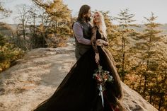 a man and woman standing next to each other on top of a cliff with trees in the background