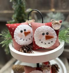 two snowmen sitting on top of a cake plate with pine cones in the background