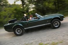 a man driving a green sports car down a road with trees in the back ground