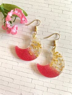 two pairs of earrings with red and gold glitters on them, sitting next to a pink flower