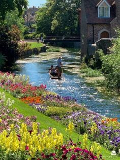 a small boat traveling down a river surrounded by lots of flowers and greenery in front of a brick building