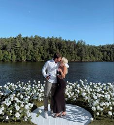 a man and woman are standing in front of flowers by the water with their arms around each other