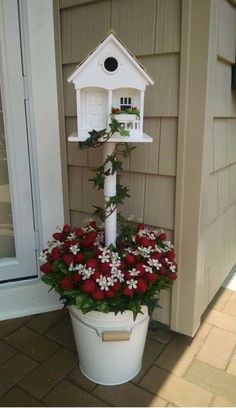 a white birdhouse sitting on top of a bucket filled with red and white flowers