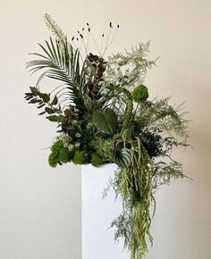 a white vase filled with lots of different types of flowers and greenery next to a wall