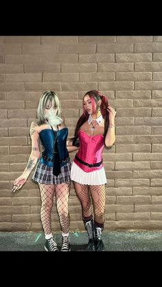 two women dressed in costumes standing next to each other near a brick wall and holding cell phones