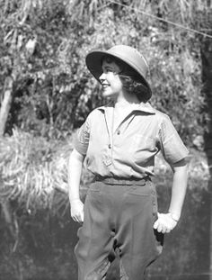 black and white photograph of a woman in overalls with hat standing on the grass