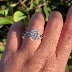 a woman's hand with a diamond ring on top of her finger, in front of some bushes