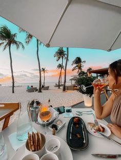 a woman sitting at a table with food and drinks in front of the ocean on a sunny day