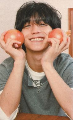 a young man holding three apples in front of his face while sitting at a table