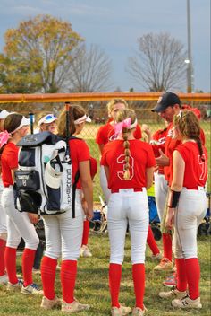 a group of girls in red and white baseball uniforms standing on the field with their backs to each other