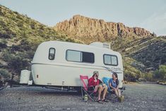 two people sitting in chairs next to an rv on the side of a road with mountains in the background