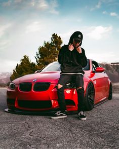 a man sitting on the hood of a red car talking on his cell phone while wearing a black hat