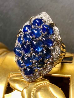 An incredibly colorful cocktail ring crafted in platinum and set with approximately 11.50cttw in vibrant royal blues cabochon sapphires surrounded by approximately .50cttw in G-H color Vs1-2 clarity round diamonds. A gorgeously made and substantial ring that you will absolutely know that you have on; it makes quite the statement! Dimensions/Weight: Ring measures 1.25" by .95" and weighs 23.8g. Size 7 (sizable). Condition: All stones are secure and in perfectly wearable condition. R-BCYYE Luxury Sapphire Cabochon Ring, Luxury Sapphire Cabochon Ring For Anniversary, Luxury Blue Cabochons For Anniversary, Luxury Blue Round Cabochons, Fine Jewelry Sapphire Cabochon Ring, Elegant Cabochon Sapphire Ring, Fine Jewelry Sapphire Ring With Cabochon Cut, Luxury Multi-stone Sapphire Gemstones, Formal Cabochon Sapphire Ring