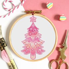 a cross stitch christmas tree on a pink background next to scissors, thread and spools