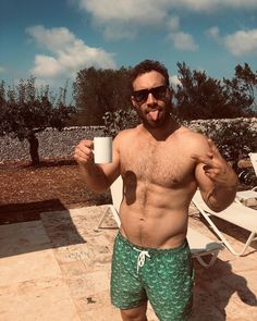 a shirtless man holding a coffee cup in his right hand while standing next to a pool