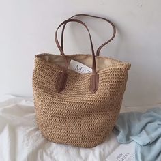 a brown straw bag sitting on top of a bed next to a book and pillow