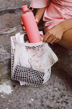 a woman sitting on the ground with a pink water bottle in her hand and a mesh bag next to her