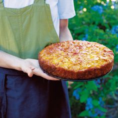 a person in an apron holding a cake on a tray with flowers behind them and bushes