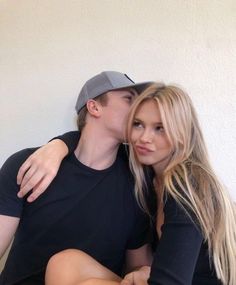 a man and woman sitting next to each other in front of a white wall kissing