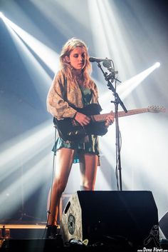 a woman standing in front of a microphone and guitar on stage with lights behind her