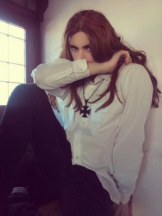 a woman with long hair sitting on the floor in front of a window wearing a white shirt and black pants