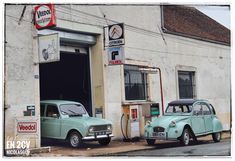 two old cars are parked in front of a gas station, one is green and the other is blue