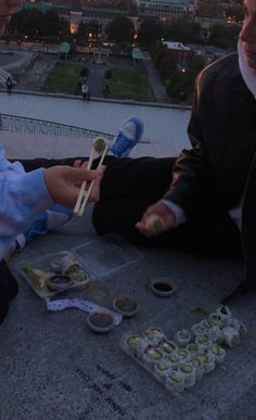 two people sitting on the ground eating sushi and chopsticks in front of a cityscape