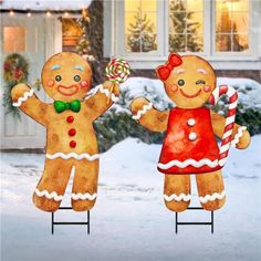 two gingerbread man and woman standing on top of a snow covered yard with candy canes