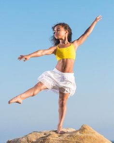 a woman in yellow shirt and white skirt standing on rock with arms out to the side