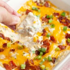 a hand holding a tortilla chip over a casserole in a white dish