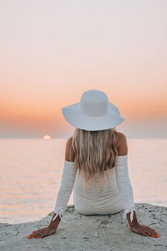 a woman sitting on top of a beach next to the ocean wearing a white hat