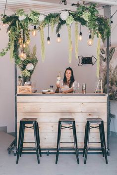 a woman sitting at a bar with three stools in front of her and flowers hanging from the ceiling