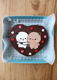 a heart shaped chocolate cake with two bears on it sitting in a blue box next to a wooden table