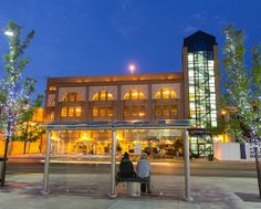 two people are sitting on a bench in front of a building with christmas lights at night