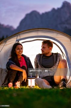 a man and woman sitting in front of a tent at night with mountains in the background