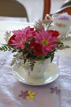 pink and red flowers are in a teacup on a white tablecloth with yellow daisies