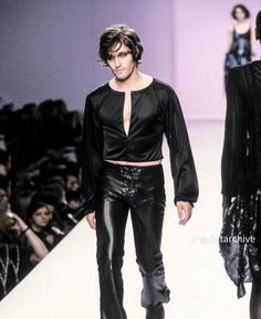 a man walking down a runway wearing leather pants and a black top with long sleeves