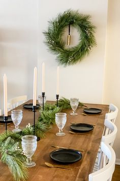 a wooden table topped with black plates and white chairs next to a wreath on the wall