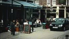 people are sitting at tables on the sidewalk in front of shops and parked cars,