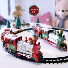 a toy train set on a table with a teddy bear and christmas tree in the background