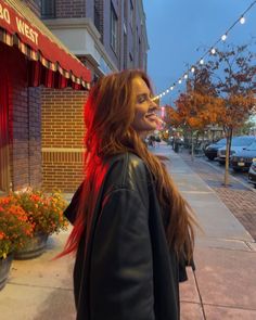 a woman with long red hair walking down the street in front of a restaurant at night