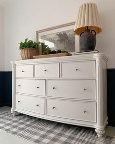 a white dresser sitting in the corner of a room next to a lamp and potted plant