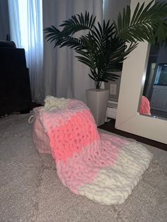 a pink and white knitted blanket sitting on the floor next to a potted plant