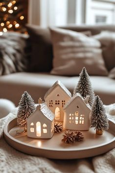 a white tray topped with small houses and pine cones on top of a couch next to a christmas tree