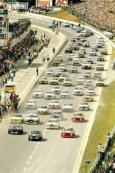 a large group of cars driving down a race track with spectators watching from the stands