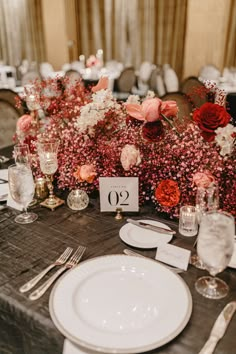 the table is set with white plates, silverware and red flower centerpieces