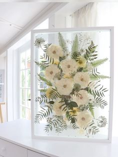 an arrangement of flowers is displayed in a frame on a white countertop next to a window