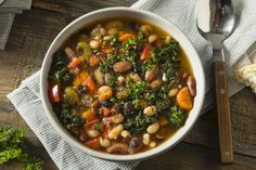 a bowl of soup with beans, kale and carrots next to a spoon